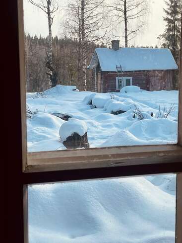 Through a window, thick snow blankets the ground. A rustic wooden cabin stands among bare trees, creating a peaceful, wintery scene in nature’s quiet embrace.