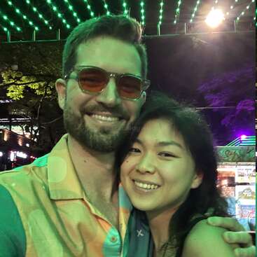 Um casal sorridente tira uma selfie sob luzes verdes e roxas vibrantes à noite, abraçando-se alegremente. O fundo sugere um local ao ar livre animado com decorações iluminadas.
