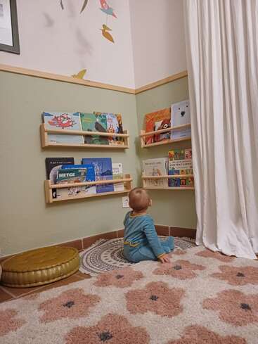 Ein Baby in einem blauen Schlafanzug sitzt auf einer kuscheligen Matte und schaut sich bunte Bücher in den niedrigen Regalen einer gemütlich eingerichteten, kinderfreundlichen Leseecke an.