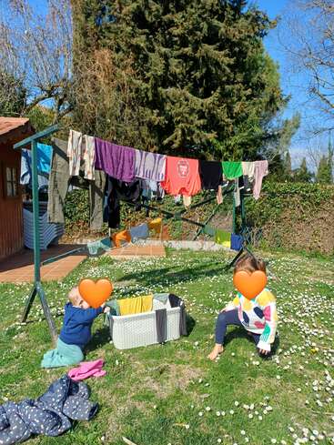 Zwei Kinder spielen auf einer Rasenfläche, die mit kleinen weißen Blumen übersät ist. In der Nähe hängt Wäsche an Wäscheleinen. Es ist ein sonniger Tag, im Hintergrund stehen hohe Bäume.