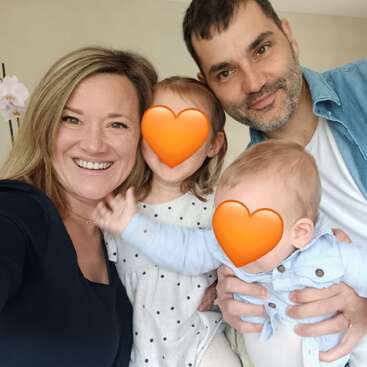 Eine glückliche vierköpfige Familie posiert für ein Selfie. Die Eltern lächeln, während die Gesichter der Kinder aus Datenschutzgründen mit orangefarbenen Herz-Emojis verdeckt sind.