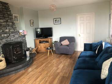 Das Bild zeigt ein gemütliches Wohnzimmer mit einem Kamin, einem Fernseher und einer blauen Couch, das mit seinem Holzboden und den hellen Wänden eine warme und einladende Atmosphäre ausstrahlt.