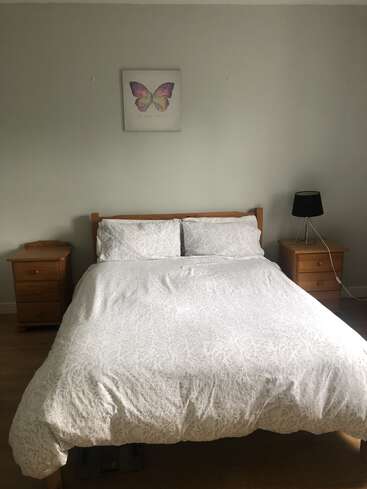 Das Bild zeigt ein ruhiges Schlafzimmer mit einem ordentlich gemachten Bett mit weißer Bettwäsche, zwei hölzernen Nachttischen und einem Schmetterlingsbild an der Wand vor einem hellen Hintergrund.