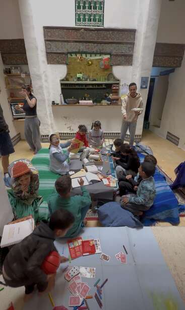 Eine Gruppe von Kindern und Erwachsenen versammelt sich in einem gemütlichen Raum, wo sie sich kreativ betätigen, zeichnen, Karten spielen und miteinander plaudern. Der Raum strahlt Wärme und Lebendigkeit aus.