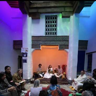 Um grupo de músicos se apresenta em um pequeno palco, numa sala com iluminação aconchegante. O público está sentado bem pertinho, assistindo. O ambiente é íntimo, colorido e acolhedor.