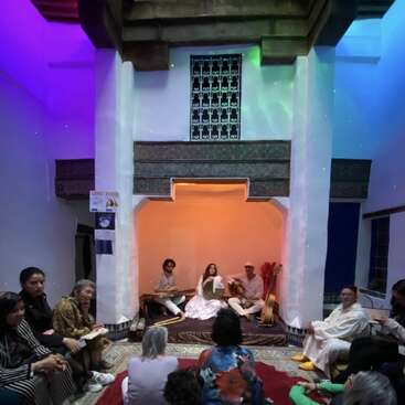 Um grupo de músicos se apresenta em um pequeno palco, numa sala com iluminação aconchegante. O público está sentado bem pertinho, assistindo. O ambiente é íntimo, colorido e acolhedor.
