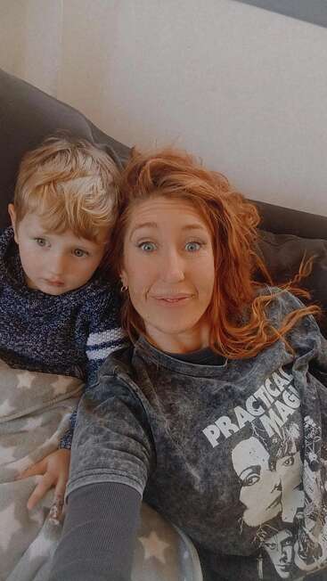 A woman with red hair and a child with blonde hair are sitting on a couch, smiling and snuggled under a star-patterned blanket, looking at camera.