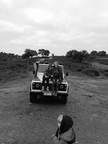 In a black-and-white photo, two people sit on a jeep’s hood in a rural landscape, while a child in a hooded jacket stands nearby. Overcast sky.