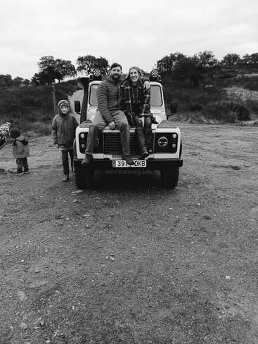 A black and white photo of four people outdoors, with a couple sitting on the hood of a jeep, children standing nearby, surrounded by trees and open space.