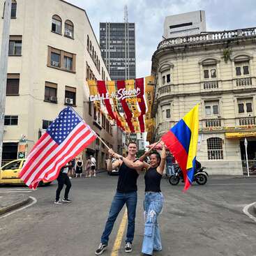 Duas pessoas agitam alegremente bandeiras dos Estados Unidos e da Colômbia em uma rua vibrante da cidade, cercada por edifícios históricos, faixas festivas no alto e alguns pedestres e veículos nas proximidades.