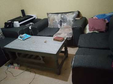 Essa imagem mostra uma sala de estar com sofás escuros, uma mesa de centro, livros e um teclado no canto. Almofadas, cabos e sapatos estão visíveis.