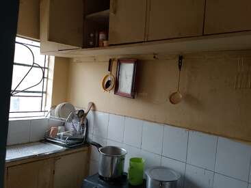 This is a small, modest kitchen with upper cabinets, hanging utensils, a cooking pot on the stove, a dish rack, and light coming through the window.