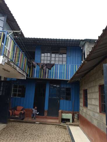 This image shows a blue two-story house with a balcony, colorful railings, some laundry drying, an armchair, a small child, and paved courtyard area.