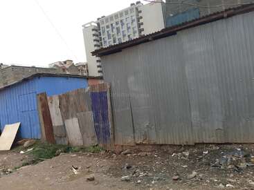 Corrugated iron sheet structures stand on a dirt lot with scattered litter, while tall modern buildings rise in the background, highlighting an urban contrast.