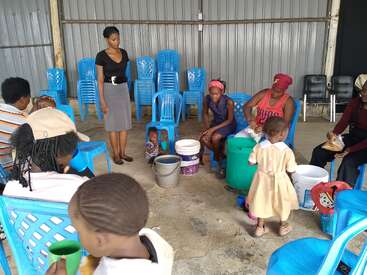 A group of women and children sit on blue plastic chairs indoors. Some are preparing food with buckets, others watch. The atmosphere seems casual and communal.