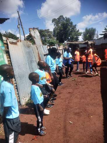 A group of children and adults, some in blue shirts, others in orange vests, stand and interact in a dirt compound with corrugated metal fences.