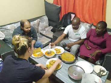 Cuatro personas están sentadas juntas en un acogedor salón, compartiendo una comida. Sobre la mesa hay platos y cuencos con comida; todos parecen felices.