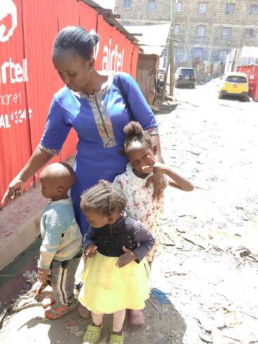Une femme vêtue d'une robe bleue se tient debout avec trois jeunes enfants dans une rue accidentée et ensoleillée, près de barrières rouges Airtel, avec des véhicules et des bâtiments en arrière-plan.