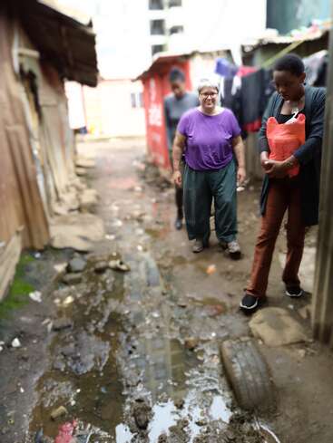 Tres personas caminan por un callejón estrecho y embarrado, flanqueado por construcciones improvisadas. Una de ellas lleva una bolsa roja. El suelo está mojado y lleno de escombros.