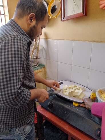 Un hombre con una camisa a cuadros está cortando tofu en cubitos en una cocina. Otra persona le ayuda sujetando la tabla de cortar con firmeza.