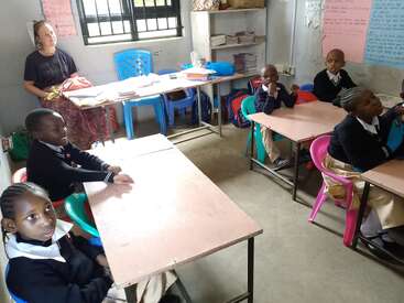 Una escena en el aula en la que se ve a seis jóvenes estudiantes con uniforme sentados en sus pupitres, una profesora al fondo, sillas de colores, carteles y material didáctico.