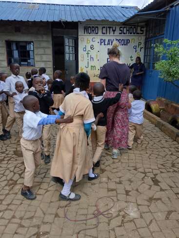 Un grupo de niños pequeños con uniforme escolar se reúne frente a «Word City Schools» junto a dos adultos. Parecen alegres, interesados y charlan animadamente en el patio.