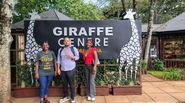 Tres personas posan sonrientes delante de un gran cartel que dice «GIRAFFE CENTRE», flanqueado por ilustraciones de jirafas. Los árboles, la vegetación y los edificios de madera crean un fondo frondoso.