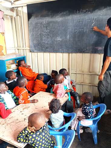 Un grupo de niños pequeños observa con atención a una profesora mientras resuelve problemas matemáticos en la pizarra, en un aula sencilla con sillas de plástico y recursos mínimos.