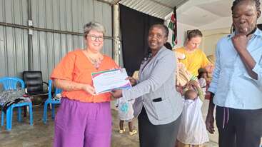 Two women stand together, smiling, as one presents a certificate to the other. Several people and children are in the background. The setting appears informal.