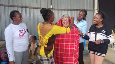 A group of five women stand together, laughing and smiling. One woman is being draped in a red checked cloth by the others. Joyful moment.