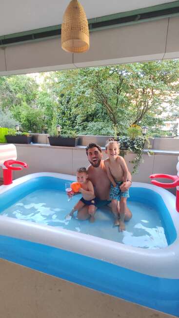 Um cara alegre e duas crianças estão brincando juntos numa pequena piscina inflável num pátio, cercado por vegetação e vasos de plantas, curtindo as diversões do verão.