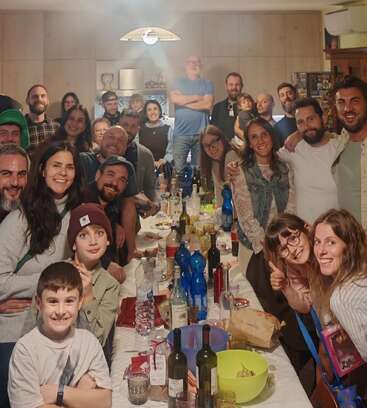 Um grande grupo de pessoas se reúne alegremente em torno de uma longa mesa cheia de comida, bebidas e garrafas, sorrindo e posando juntos em um ambiente aconchegante.