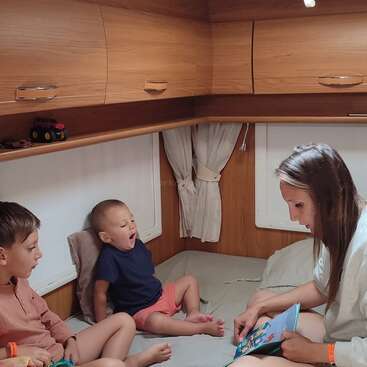 Una mujer lee un libro a dos niños pequeños dentro de una acogedora caravana. Uno de los niños bosteza, mientras que el otro escucha con atención, rodeado de armarios de madera y una suave iluminación.