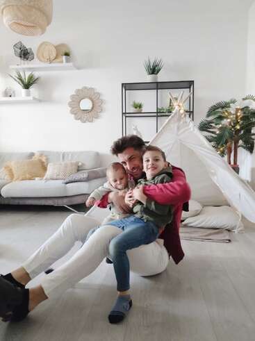 Un père joyeux serre ses deux jeunes fils dans ses bras, assis par terre dans un salon moderne et confortable, décoré de manière ludique, avec notamment un tipi et des plantes d'intérieur.