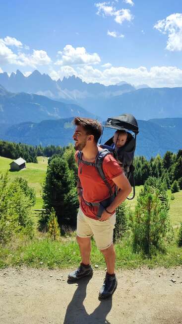Un homme portant un bébé dans un sac à dos de randonnée traverse un paysage montagneux pittoresque avec des arbres verdoyants, un ciel bleu et des sommets escarpés au loin.
