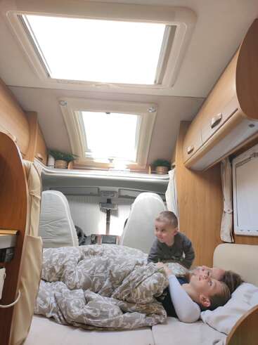 Un intérieur de camping-car confortable avec des armoires en bois et de grandes lucarnes. Deux enfants et un adulte se détendent sur un lit recouvert de couvertures à motifs, baignés d'une douce lumière naturelle.