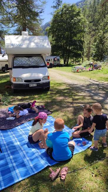 Des enfants se détendent sur des couvertures de pique-nique à l'ombre près d'un camping-car dans un camping boisé, profitant des jeux et de la nature par une journée ensoleillée.