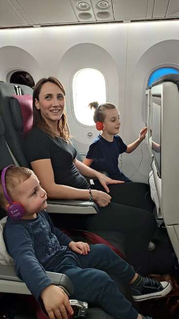 Une femme et deux enfants, équipés d'écouteurs, sont assis dans un avion. Les enfants sont absorbés par leurs écrans. Tout le monde semble détendu et heureux.