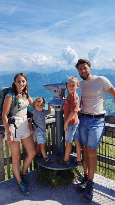 Une famille de quatre personnes souriantes pose devant un télescope panoramique sur une plate-forme en bois, entourée de montagnes, d'un ciel bleu et de nuages par une journée ensoleillée.