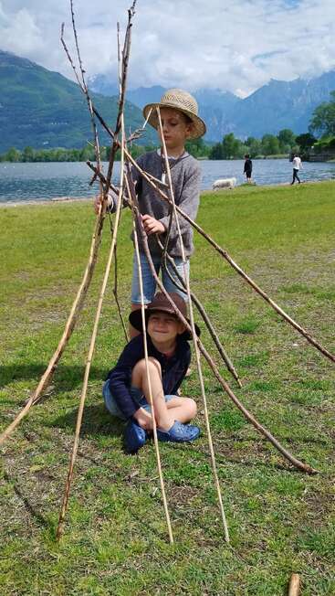 Deux enfants, coiffés de chapeaux, construisent une petite structure en forme de tipi avec de longs bâtons au bord d'un lac. Les montagnes et les nuages forment un décor naturel pittoresque avec de la verdure.