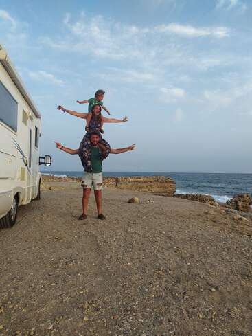 Trois personnes forment une tour humaine ludique près d'un camping-car au bord de la mer, chacune écartant les bras, profitant d'une belle journée ensoleillée sous un ciel bleu.