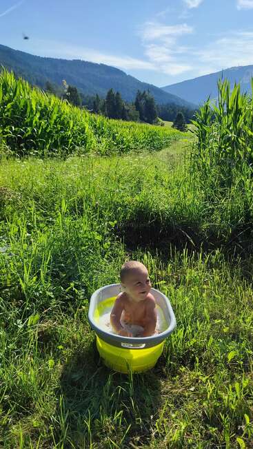 Un bébé souriant est assis dans une baignoire jaune remplie d'eau, au milieu d'une pelouse verdoyante, entouré de grands champs de maïs et de montagnes, sous un ciel bleu éclatant.