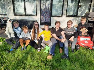 Seven children sit on green grass in front of a house, smiling happily. Two hold plush toys and one sits on a red toy car. Sunny day.