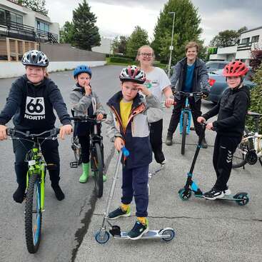 Seis crianças, usando capacetes, estão reunidas em uma rua do subúrbio com bicicletas e scooters. Elas parecem alegres e prontas para se divertirem juntas em uma aventura ao ar livre.
