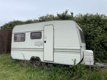 Uma pequena caravana branca vintage está estacionada na grama alta, ao lado de uma sebe alta. As janelas estão fechadas, e a cena parece tranquila e silenciosa.