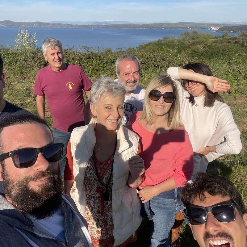 Um grupo de sete pessoas sorrindo ao ar livre, aproveitando um dia ensolarado perto de um lago, com colinas verdes ao fundo. Óculos escuros e roupas casuais.