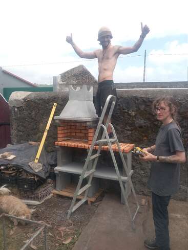 Dos personas están construyendo una barbacoa de ladrillo al aire libre. Uno está sin camiseta, de pie en una escalera dando pulgares hacia arriba, mientras que el otro sostiene herramientas y sonríe cerca. Hay un perro presente.