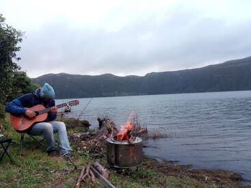 Une personne coiffée d'un chapeau bleu joue de la guitare près d'un feu de camp au bord d'un lac paisible, entourée de montagnes sous un ciel nuageux, évoquant une solitude sereine.
