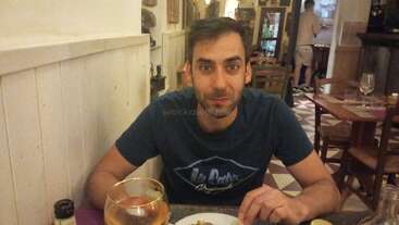 Un hombre de pelo corto y oscuro y barba está sentado en la mesa de un restaurante, con los cubiertos en la mano y una bebida y comida delante de él. El ambiente es acogedor.