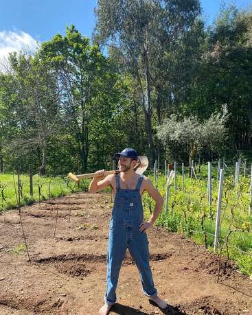 Um homem está descalço num jardim, vestindo um macacão jeans e um boné, com uma enxada pendurada no ombro, sorrindo num dia ensolarado. Ele está cercado por árvores.